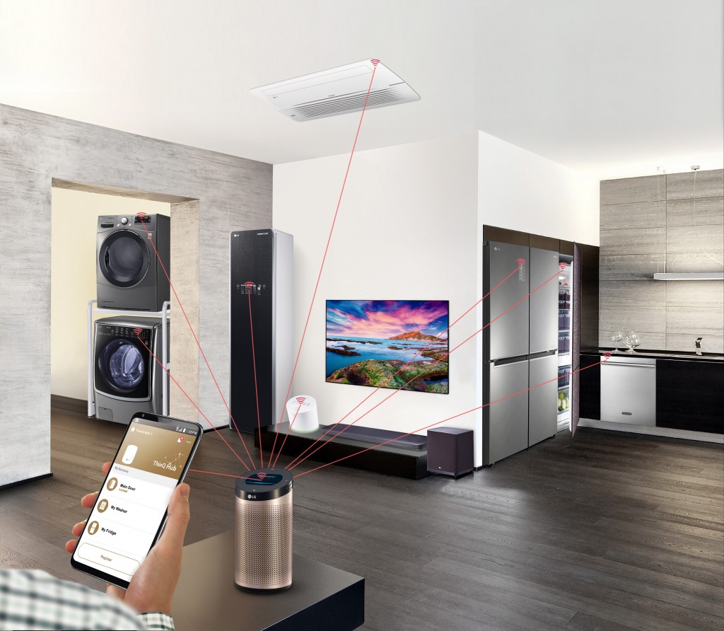 Hand holding smartphone in an apartment with lines connecting an LG ThinQ Hub speaker to a washing machine, dryer, Styler, refrigerator, dishwasher, air conditioner and air purifier