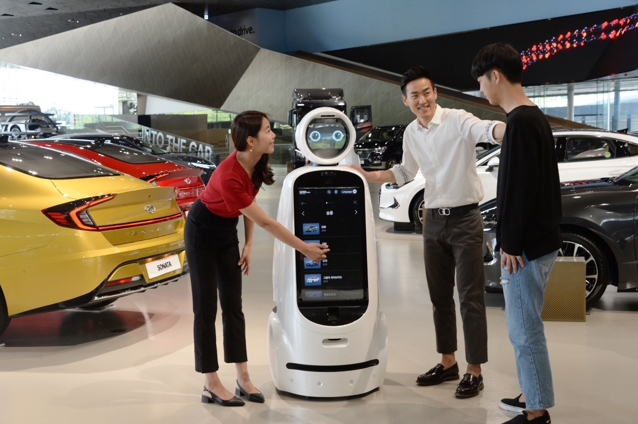 Three visitors interact with LG CLOi GuideBot in front of vehicles at Hyundai Motor Studio Goyang.