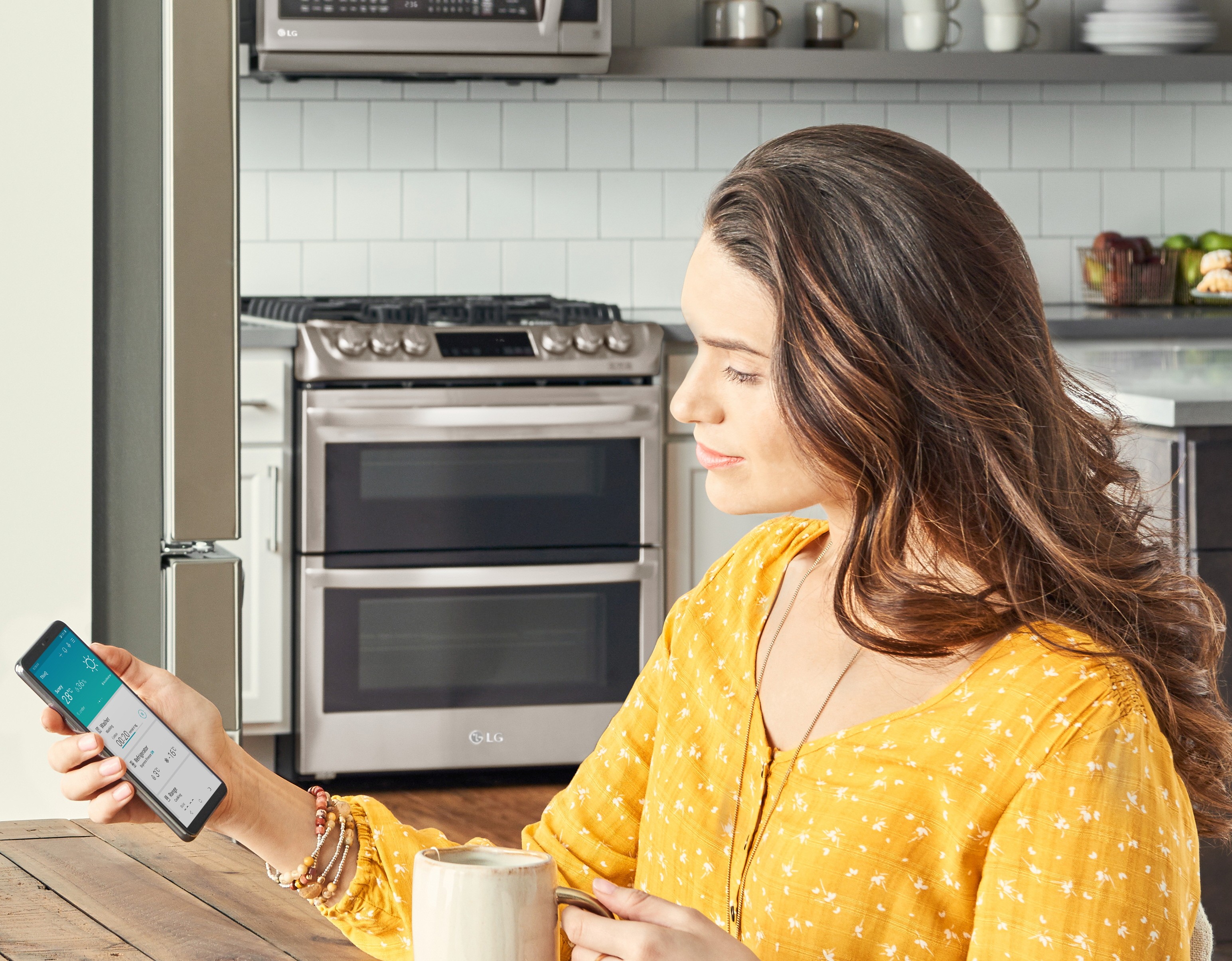 Woman sitting in her kitchen looking at a phone displaying the ThinQ™ app screen showing the device status of the washer, refrigerator and range