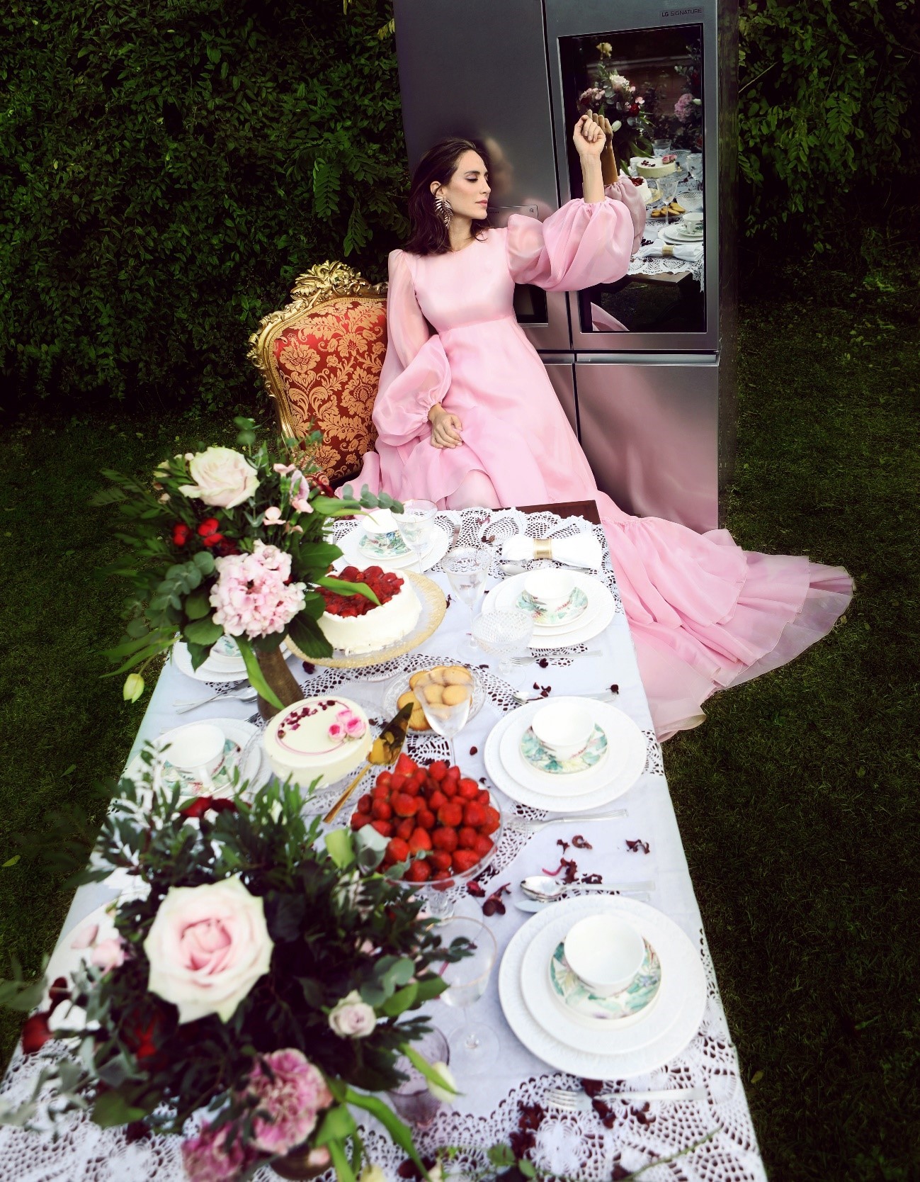 A photo recreating the Mad Hatter tea party scene from Alice in Wonderland, with fashion designer Tamara Falcó sitting at the head of a dinner table as she leans back against the LG SIGNATURE Door-in-Door refrigerator and touches the InstaView panel.