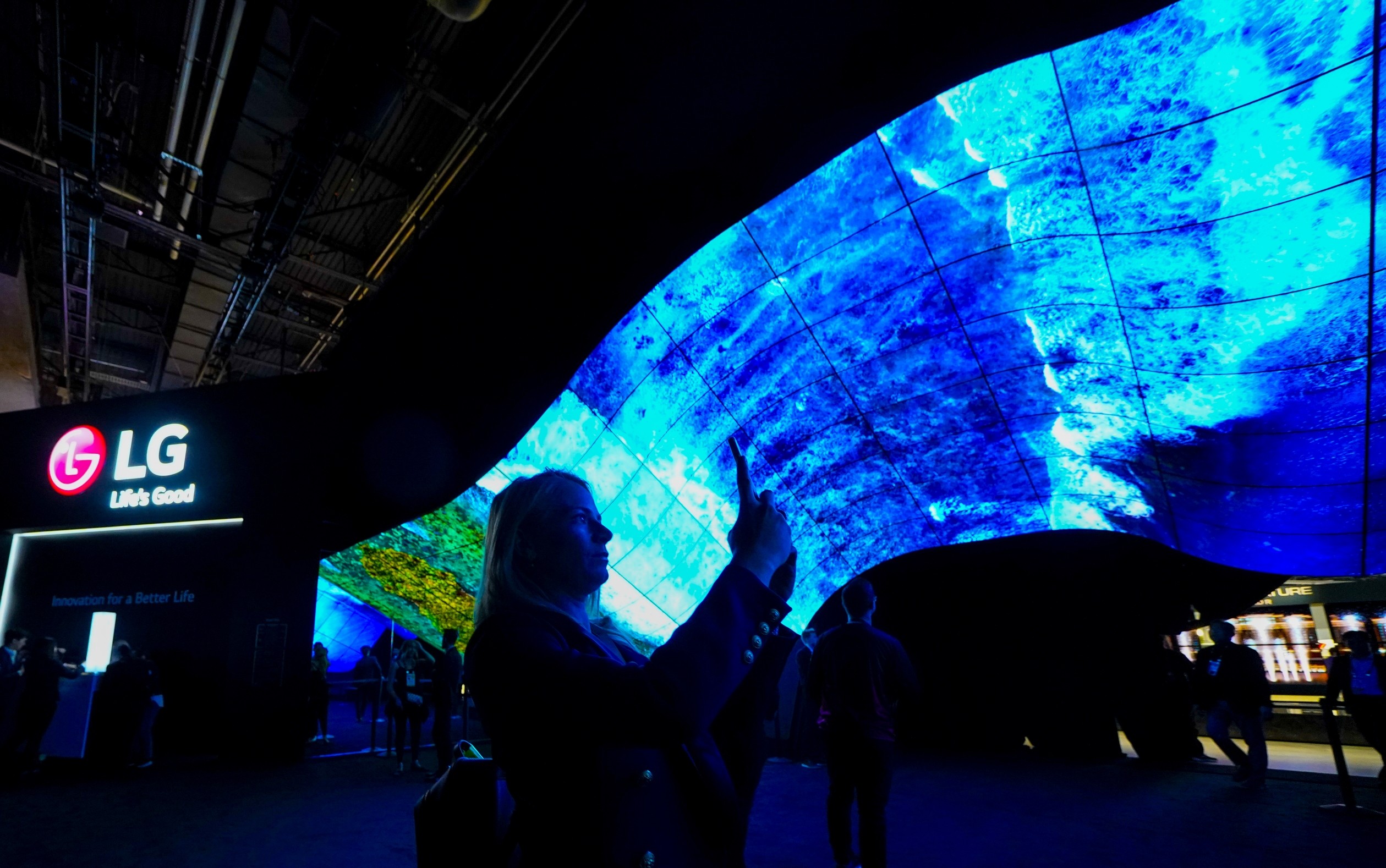 A woman taking a photo of the mesmerizing LG OLED Wave at CES 2020 as it displays vivid blue waves crashing against the coastline.