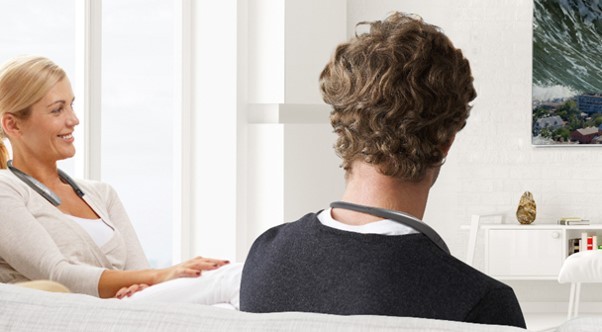 A man and a woman sit on a couch and talk to each other while wearing the LG TONE Studio Bluetooth wearable speaker.