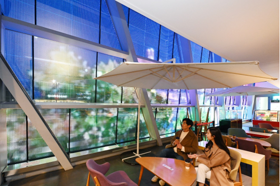 LG'nin Şeffaf LED Filmi, bir erkek ve kadın binanın içinden izlerken bir binanın yan tarafındaki renkli ağaçları görüntülüyor. Busan Sinema Merkezi