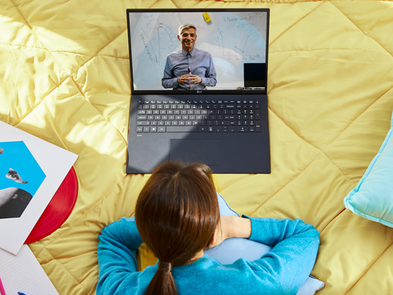 A woman relaxes on her bed while using her LG gram to take a lesson from a teacher online.