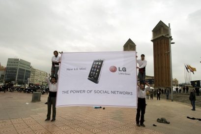 Two people stand on the shoulders of two others to display a LG Mini promotional banner next to the Venetian Towers at Plaza España, Barcelona.