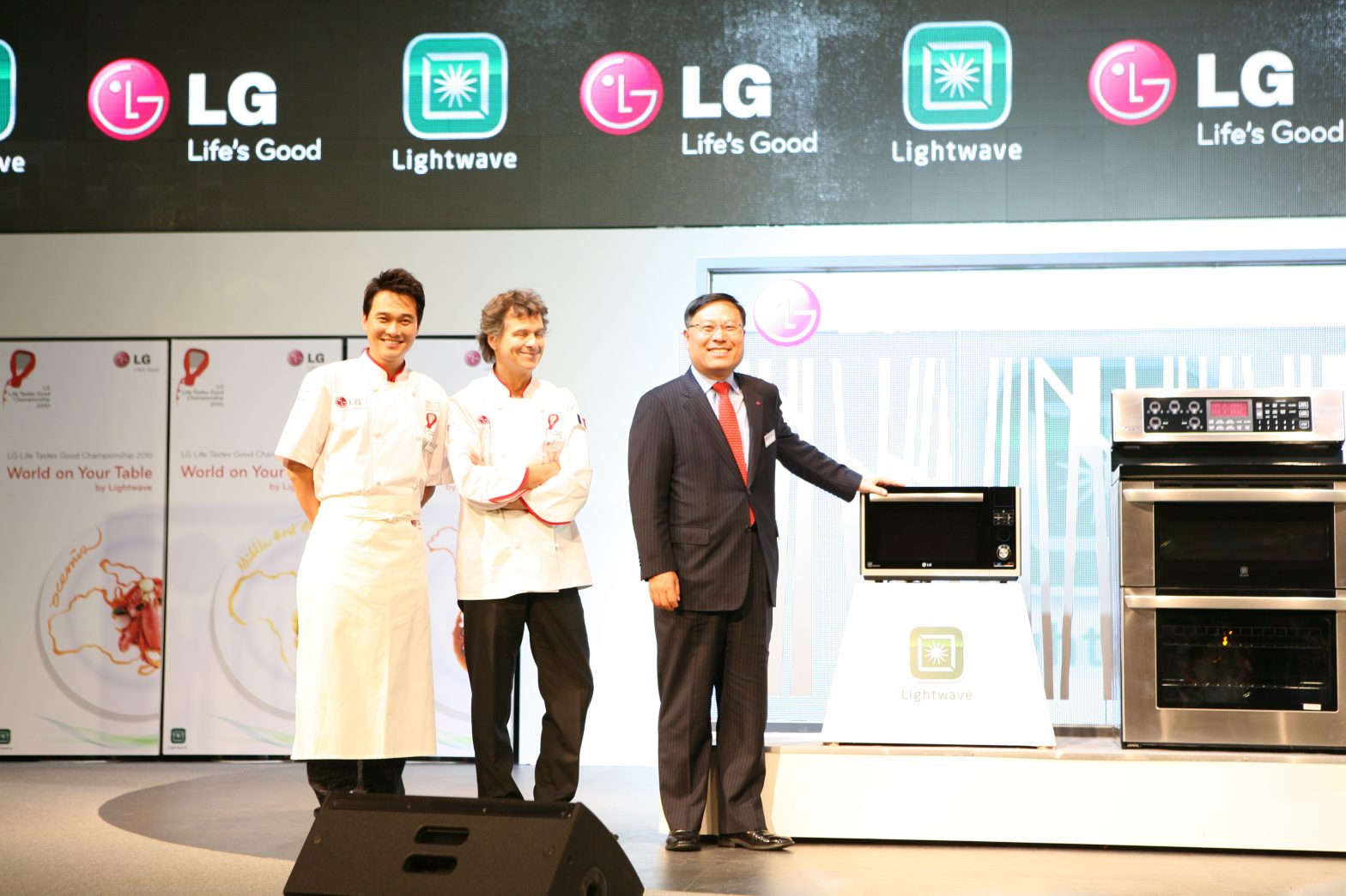 Two chefs and one LG executive pose at the front of the stage with the LG microwave and cooking oven