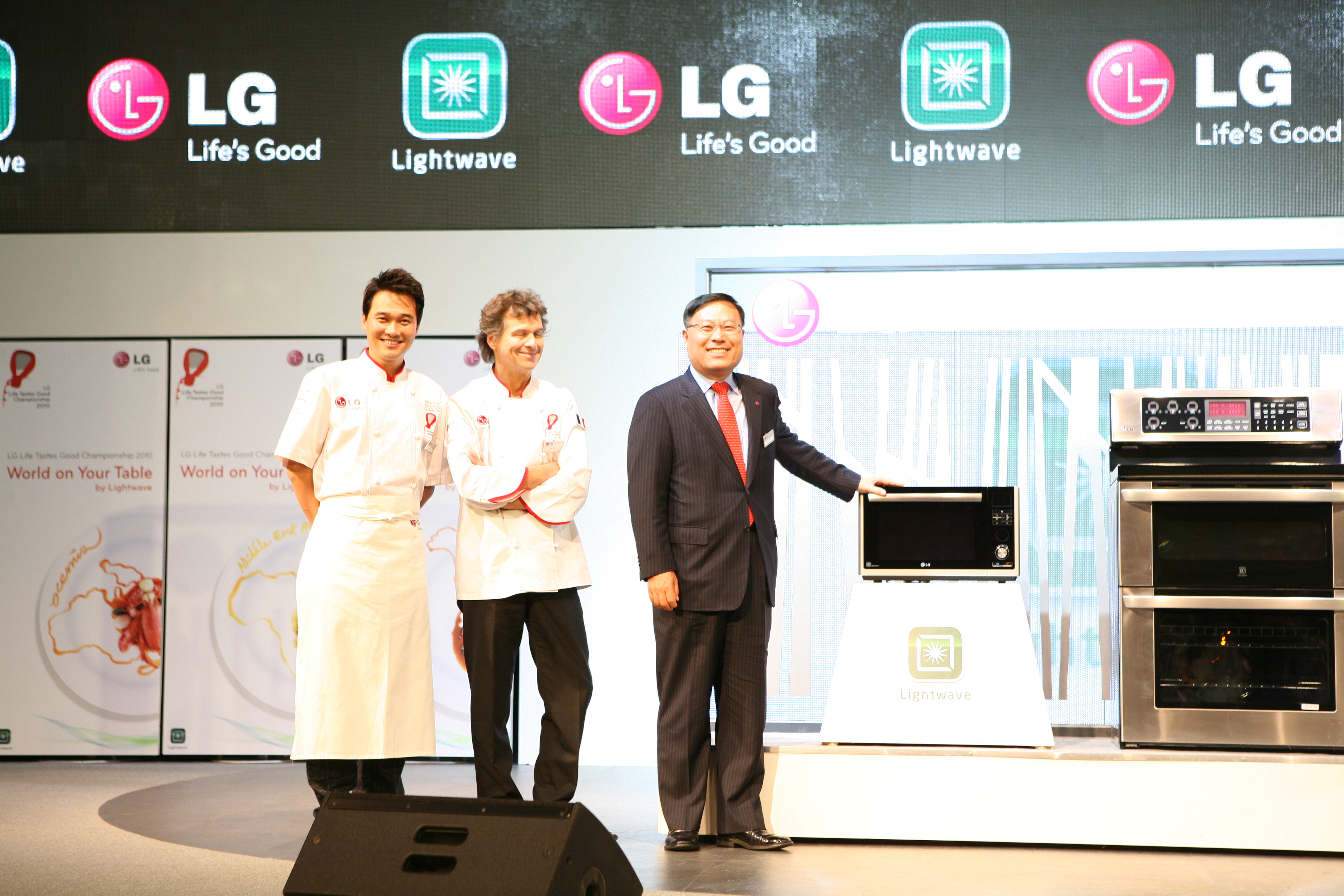 Two chefs and one LG executive pose at the front of the stage with the LG microwave and cooking oven