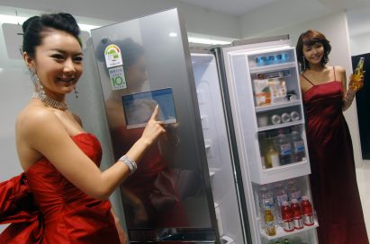 Two models pose with the open LG DIOS smart refrigerator, with one pointing at the refrigerator’s front door display