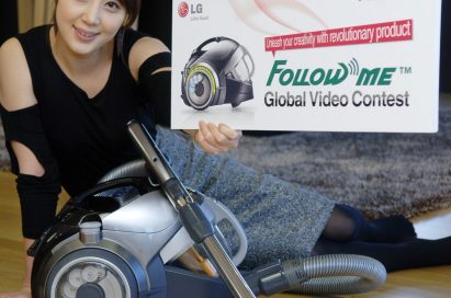 A woman sitting down on the floor holding the banner that reads ‘Follow Me Global Video Contest’ with the LG KOMPRESSOR FOLLOW ME™ vacuum cleaner
