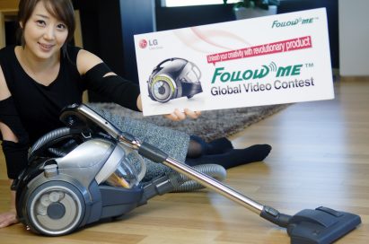 A woman sitting on the floor while posing with the banner of ‘Follow Me Global Video Contest’ in front of the LG KOMPRESSOR FOLLOW ME™ vacuum cleaner