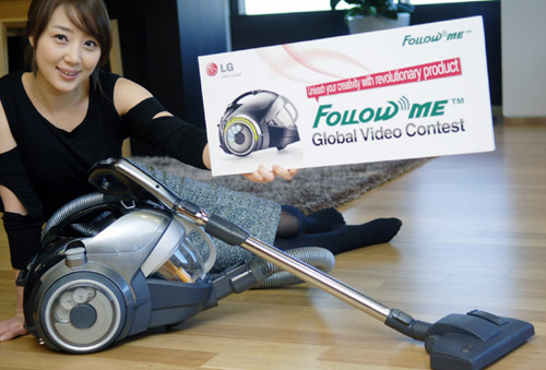A woman sitting on the floor while posing with the banner of ‘Follow Me Global Video Contest’ in front of the LG KOMPRESSOR FOLLOW ME™ vacuum cleaner