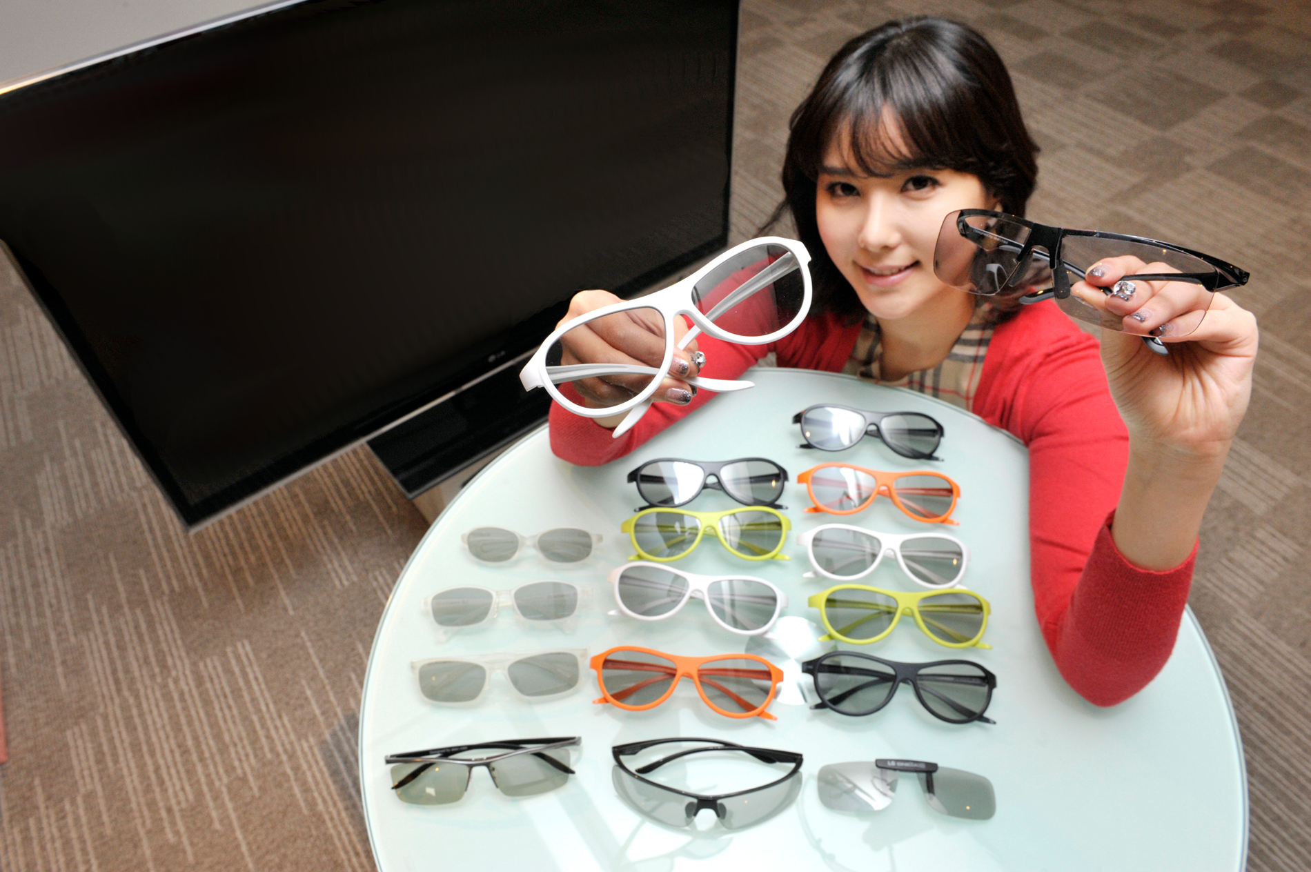 A model presenting the various types and colors of LG’s new 3D glasses lineup on a table, with the LG CINEMA 3D TV just behind her
