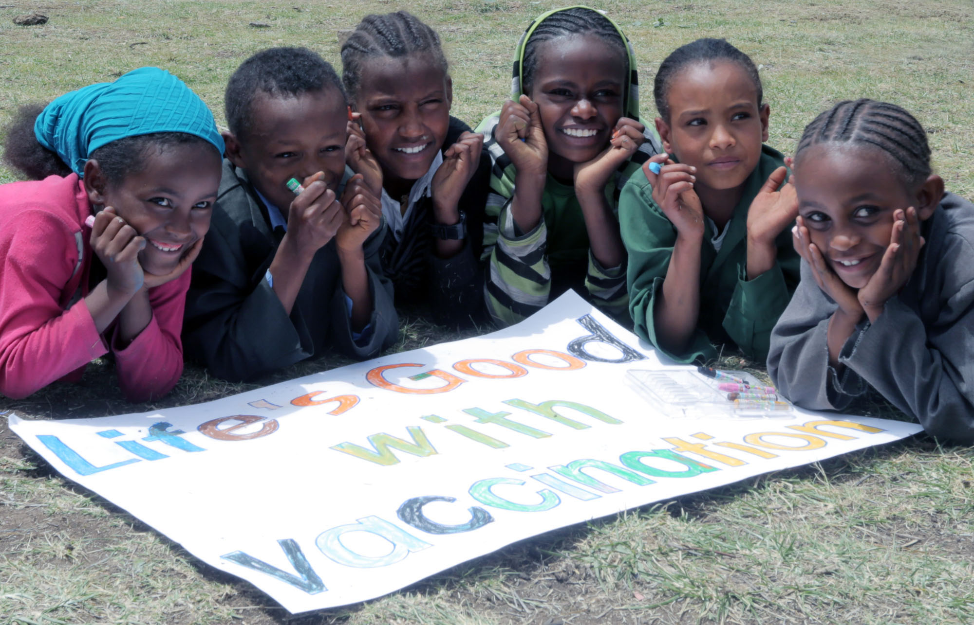 Children color a "Life's Good with vaccination" sign with crayons on the grass