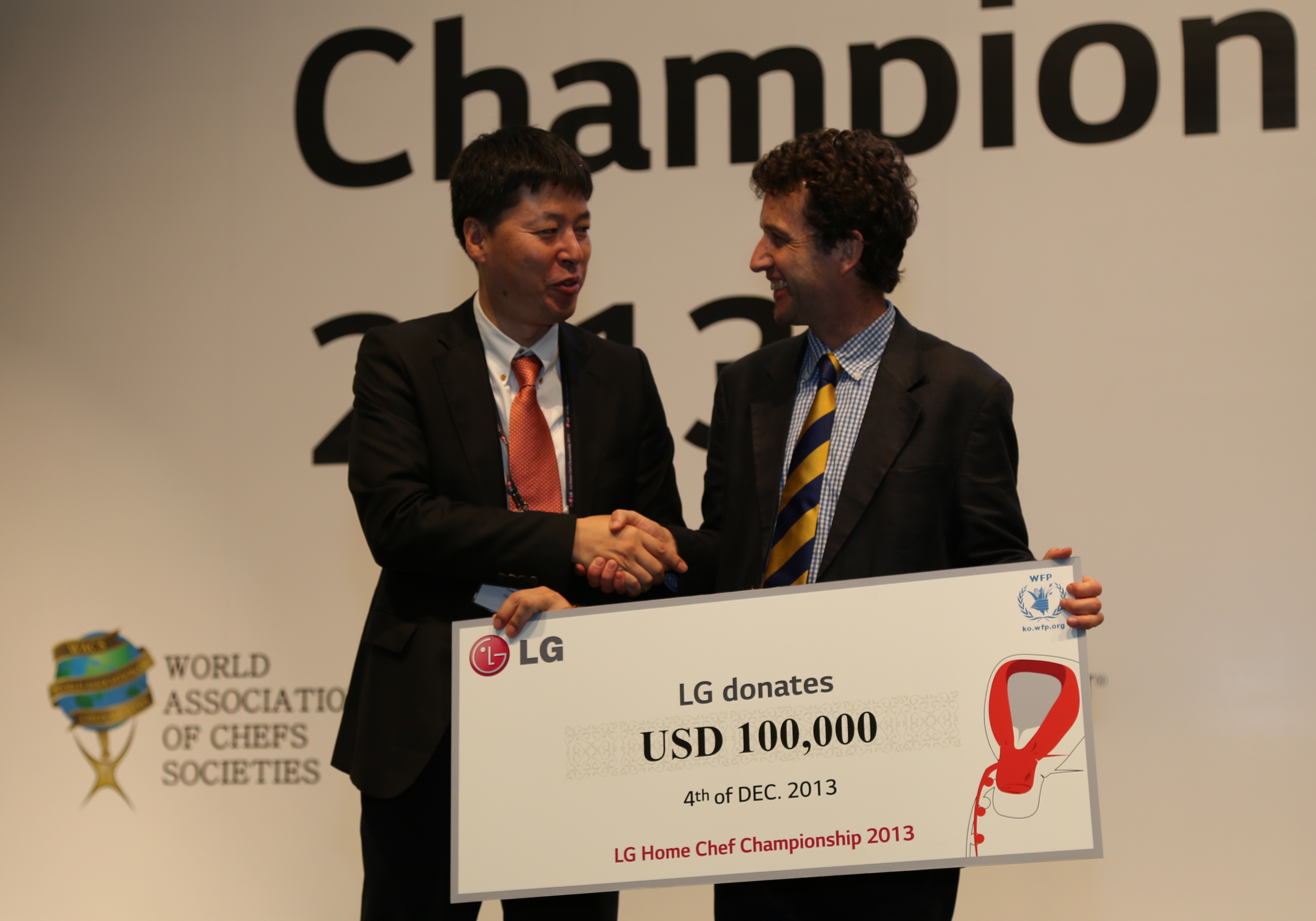 An LG representative shakes hands with a gentleman from the United Nations World Food Programme (WFP) while holding a panel showing LG’s donation of 100,00 USD at the LG Home Chef Championship 2013