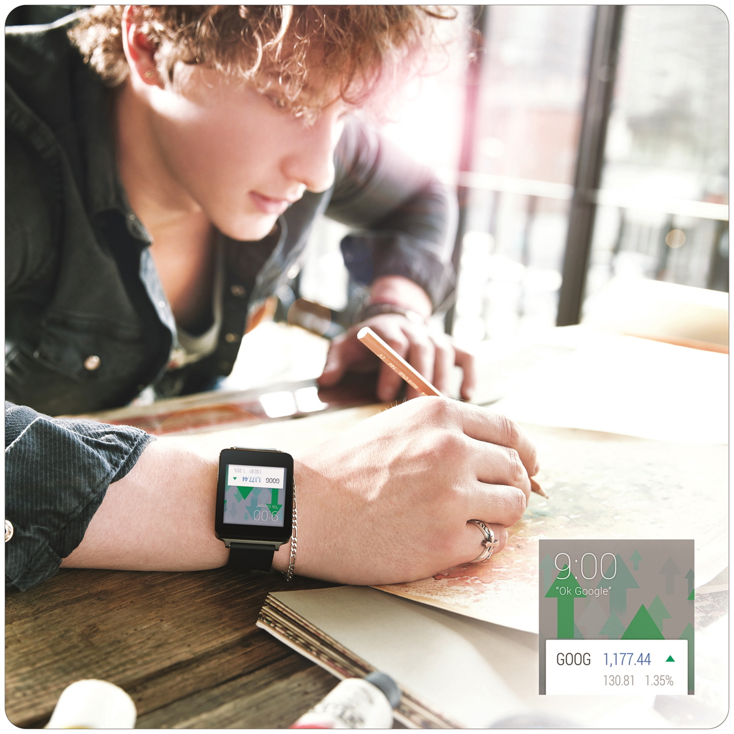 A man writes on paper while wearing the LG G Watch in Titan Black, with a small screenshot close-up of the watch’s display showing Google’s stock activity overlapping.