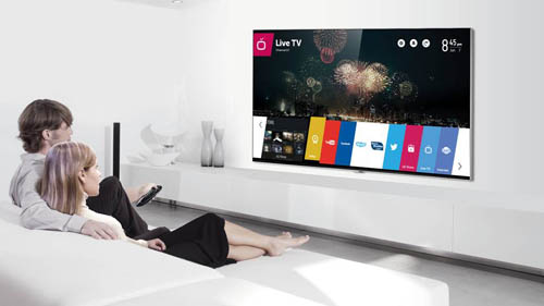 A man and woman sit on their sofa while selecting apps run by webOS with a remote on the LG Smart+ TV.