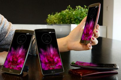 A model is putting her arm down on a table holing a LG G Flex2. And two LG G Flex2s are standing on the table showing their fronts and other twos are lying on the table – each showing front and back.