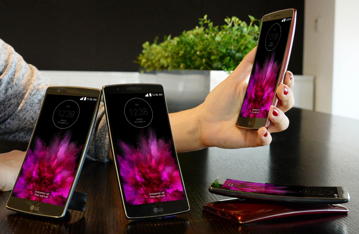 A model is putting her arm down on a table holing a LG G Flex2. And two LG G Flex2s are standing on the table showing their fronts and other twos are lying on the table – each showing front and back.