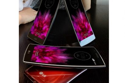 Four LG G Flex 2 smartphones in each color variant are placed on the table and a female model poses behind those sample phones.