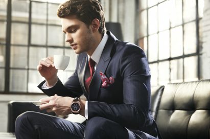 A business man in a suit wears LG's Watch Urban while drinking coffee on a couch.