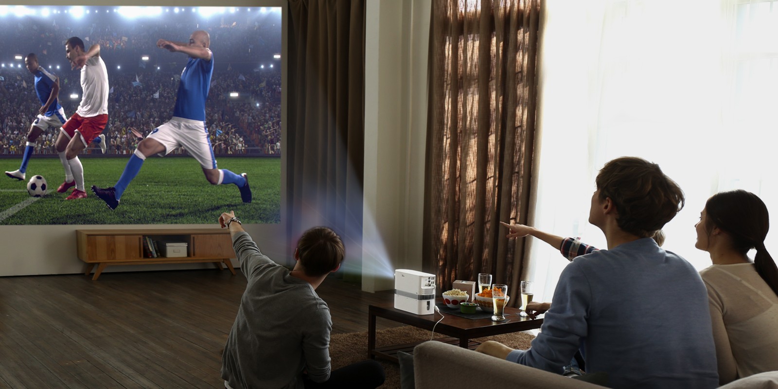 Three people watch a soccer game projected on the wall by the LG Probeam Laser Projector (model HF80J)