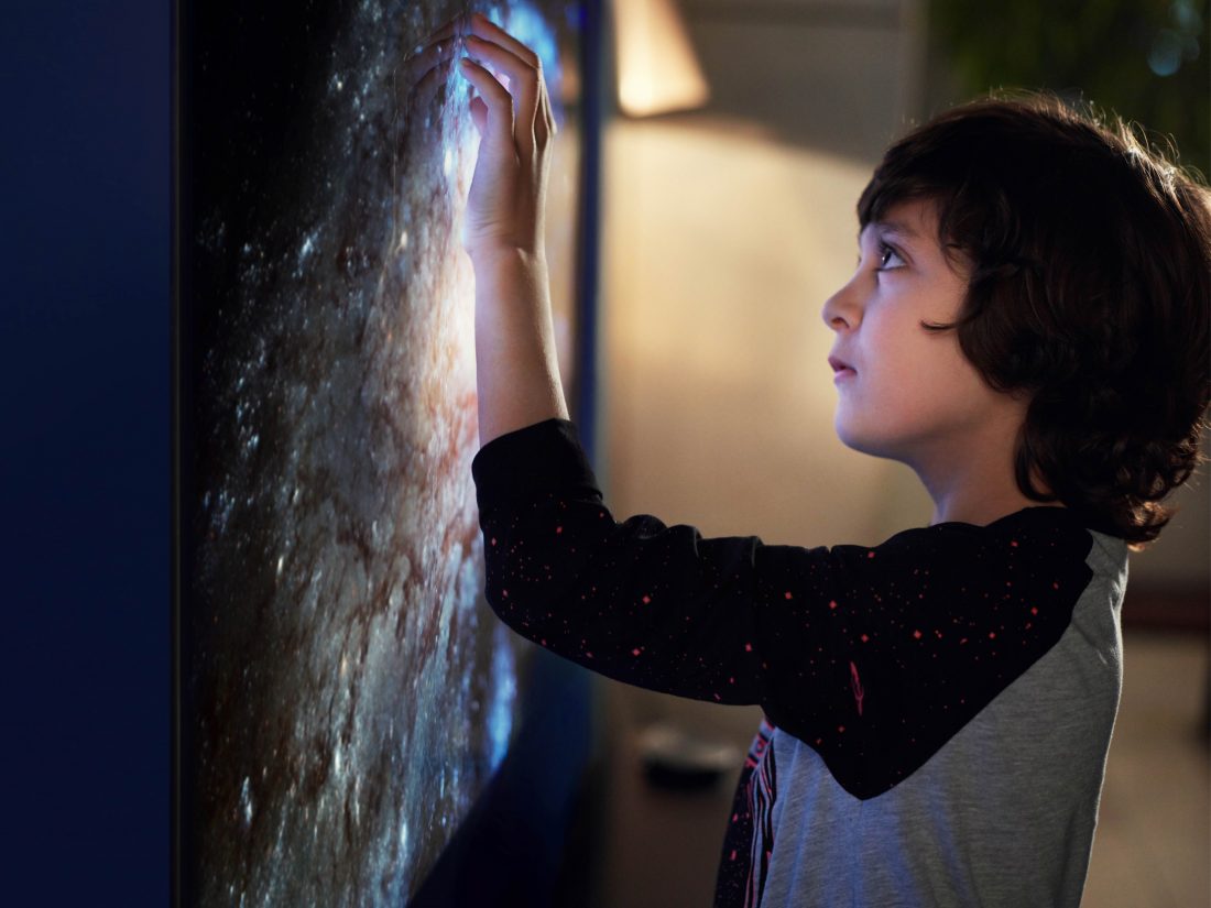 A small boy touches the screen of the LG SIGNATURE OLED TV W, there is a picture of a galaxy on the screen.