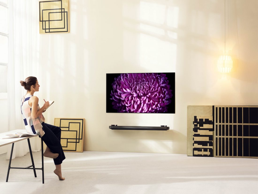 A woman rests against the table looking at an LG SIGNATURE OLED TV W (model W7) mounted on the wall.