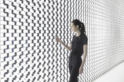 A wider angle of a woman touching one of the thousands of bright lights found on the back wall of Tokujin Yoshioka’s art exhibition.