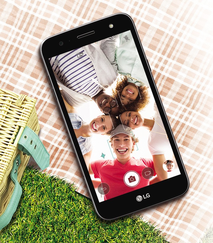 The LG Xpower2 lying on top of a picnic blanket, with a group of people captured in the camera frame while taking a selfie