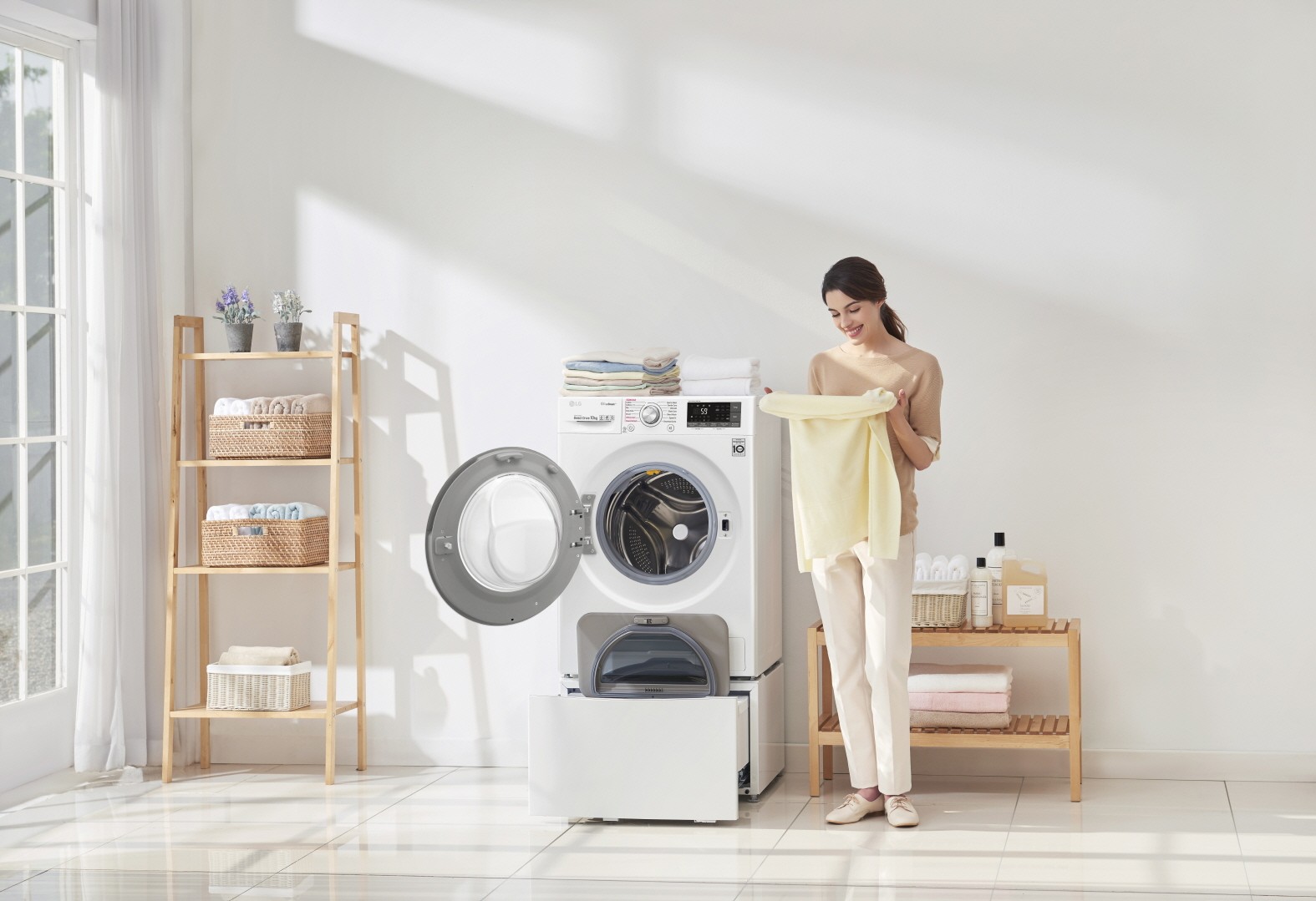 A woman holding yellow towel and standing next to LG TWINWash™ washing machine with both doors open and folded towels stacked on top of the washer in a laundry room