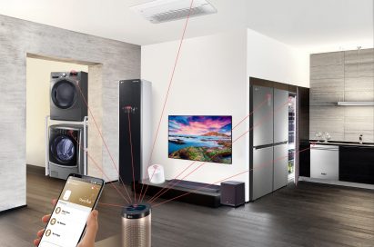 Hand holding smartphone in an apartment with lines connecting an LG ThinQ Hub speaker to a washing machine, dryer, Styler, refrigerator, dishwasher, air conditioner and air purifier