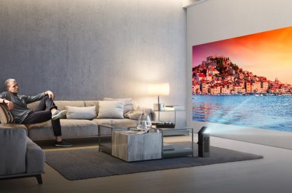 A man seated on a couch watches video projected onto a wall from LG’s HU80K series 4K UHD projector