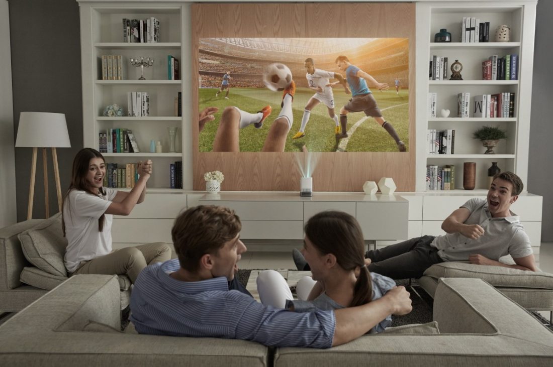 Four people sitting on couches enjoy watching a soccer game projected on the wall by the LG ProBeam Projector HF85J.