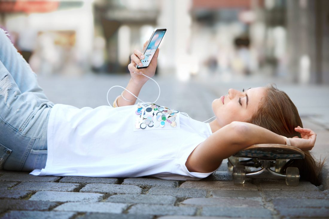 A woman lies on the ground with her skateboard while listening to music on headphones on her LG Q6
