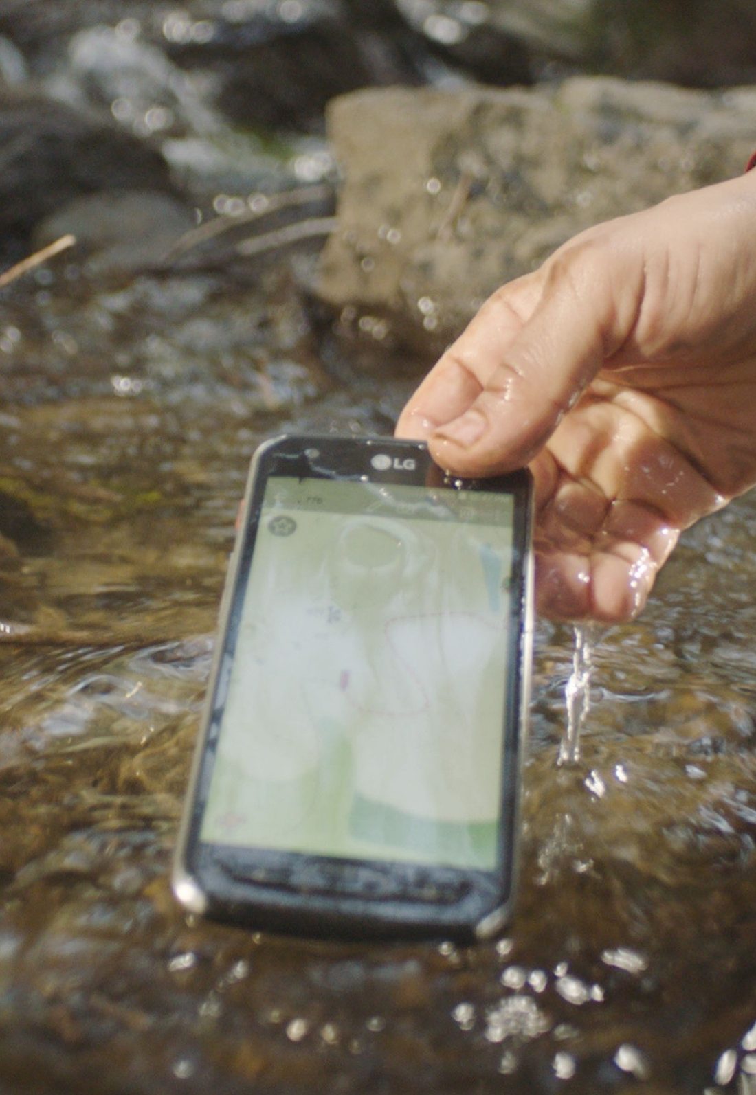 A hand lifting the water-resistant LG X Venture smartphone from water, the phone is still turned on
