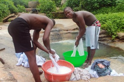 Two men hand-wash clothes in the laundry bowl by the valley stream.