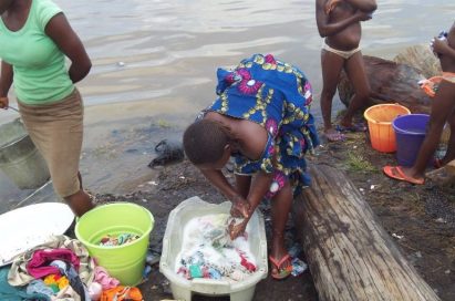 Women and children hand-wash clothes in a laundry bowl by the riverside.