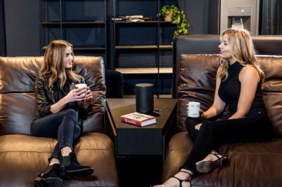 Two women sit on Natuzzi’s Colosseo sofa, talking to each other with LG’s AI smart speaker placed between them.