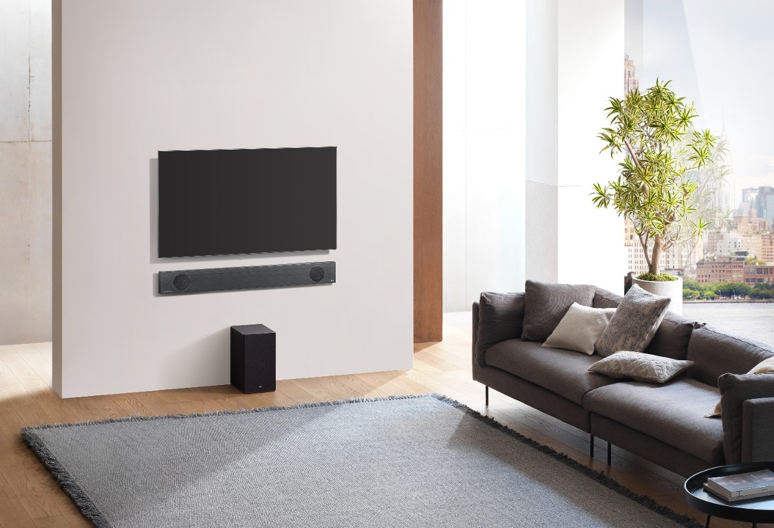 An upper view of the LG Soundbar model SL9YG installed on the wall below an LG TV, with the LG Wireless Rear Speaker Kit model SPK 8 on the floor below