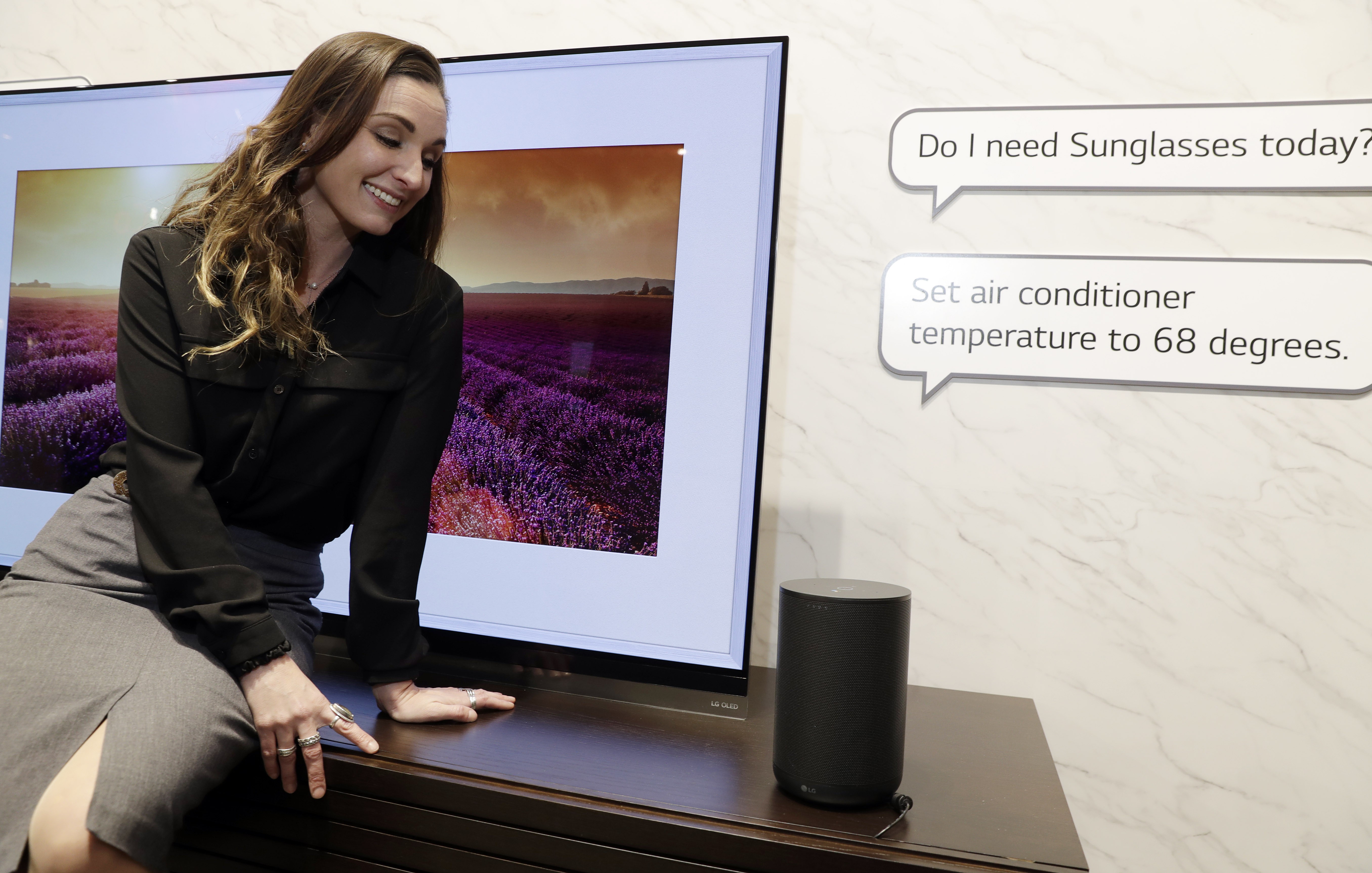 Woman sitting in front of an LG TV while facing the LG AI speaker and asking, “Do I need Sunglasses today?” and, “Set air conditioner temperature to 68 degrees”