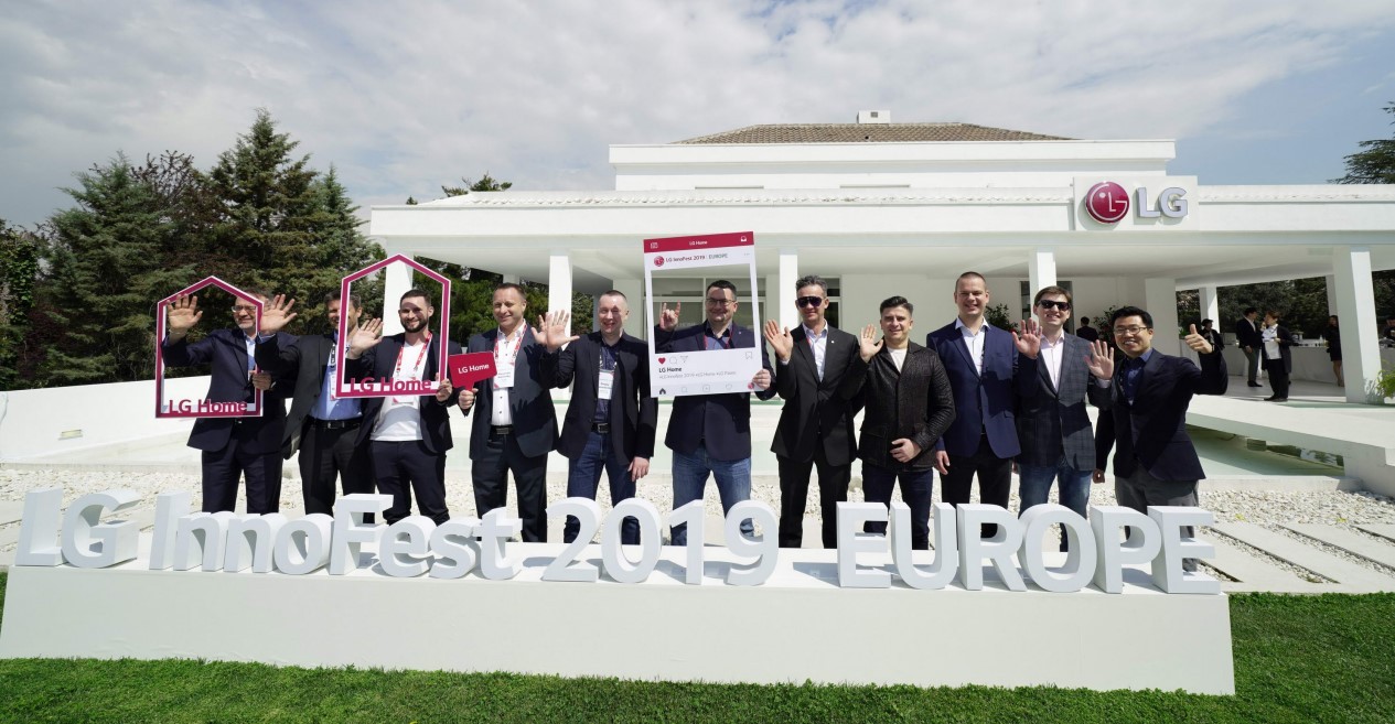 Attendees of LG InnoFest 2019 Europe wave their hands towards the camera in front of LG Home Madrid.