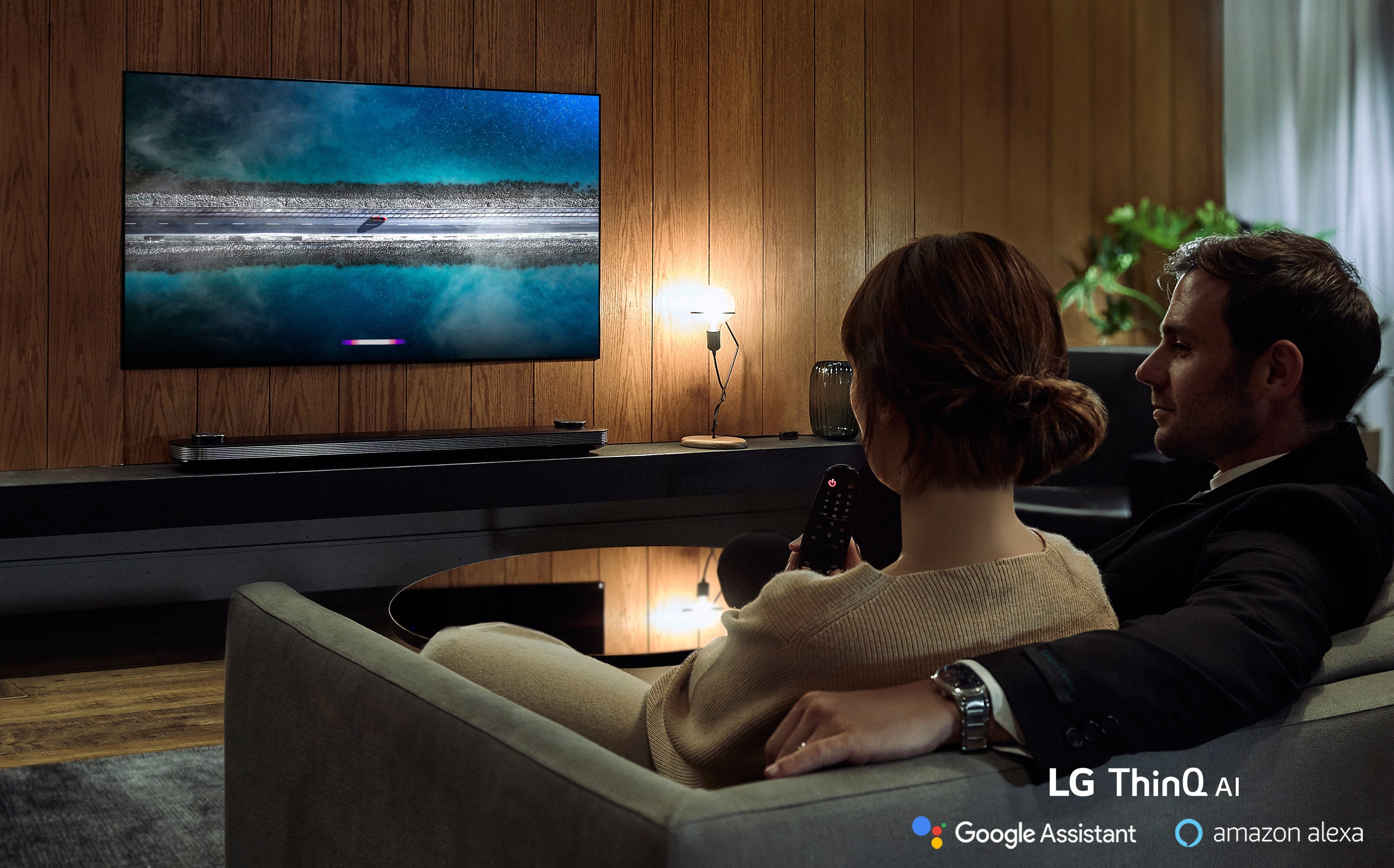 A couple is watching an LG TV supporting Amazon Alexa while the woman holds the remote in one hand