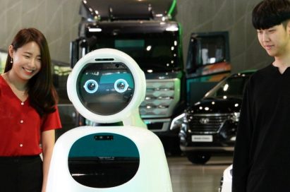 A woman and a man stand next to LG CLOi GuideBot in front of vehicles at Hyundai Motor Studio Goyang.