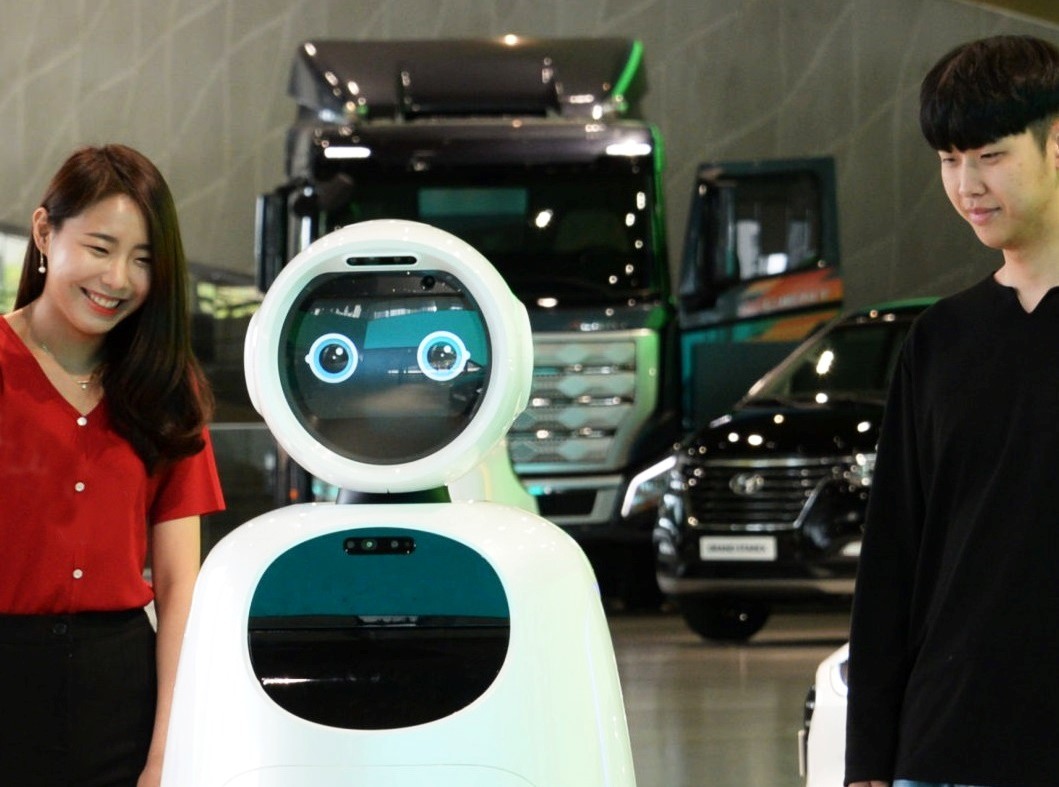 A woman and a man stand next to LG CLOi GuideBot in front of vehicles at Hyundai Motor Studio Goyang.