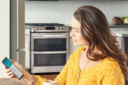 Woman sitting in her kitchen looking at a phone displaying the ThinQ™ app screen showing the device status of the washer, refrigerator and range