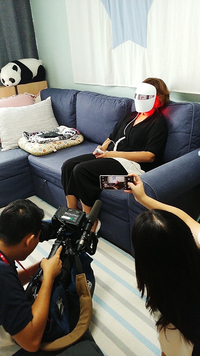 A cameraman shoots a video of a consumer sitting on the couch while wearing the LG Pra.L Derma LED Mask on her face.