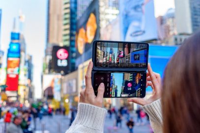 A woman holds LG G8XThinQ with LG Dual Screen in her two hands vertically to check the weather on the screen at an urban space with buildings.