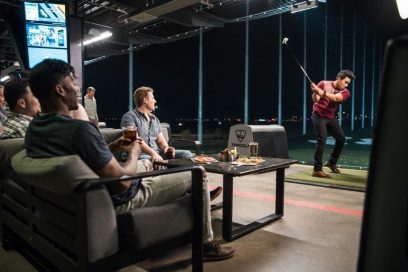 A golfer hits a tee shot as friends watch while sat on the couch at a Topgolf venue.