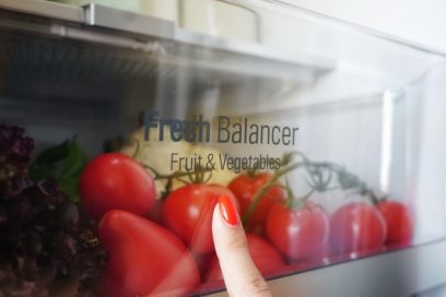 A close-up of the LG V+ refrigerator’s Fresh Balancer drawer full of tomatoes and lettuce, and a woman pressing her finger up against it.