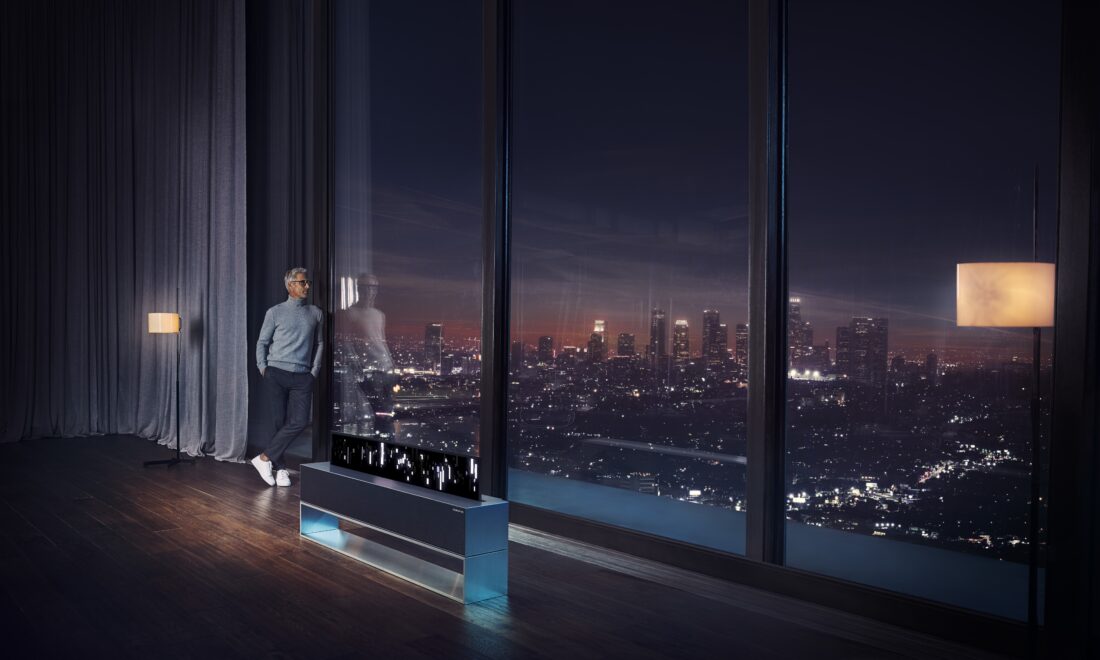 A man looking out the window at the city skyline at night, an LG SIGNATURE OLED TV R is reflecting a picture of the skyline